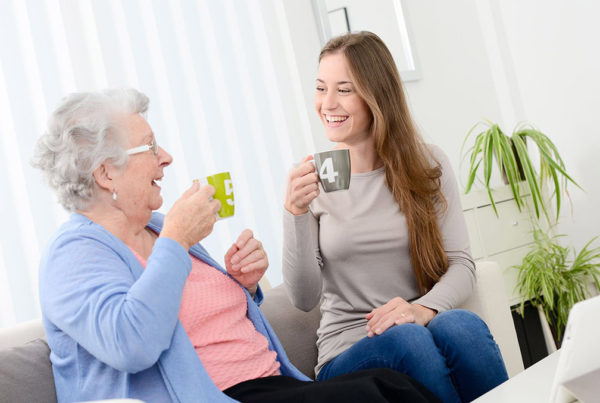 weibliche Pflegekraft und ältere Dame sitzen lachend auf der Couch mit zwei Tassen in der Hand.