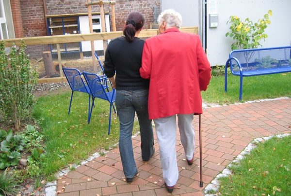 ältere Frau mit Gehstock, die sich bei einer jüngeren Frau unterm Arm eingehakt hat. Beide gehen auf einem Weg im Garten.