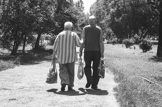 schwarz-weiß Foto von zwei älteren Herren, die auf einem Feldweg gehen. Der Rechte hat zwei Einkaufstüten in der Hand, der Linke hat in einer Hand eine Einkaufstüte und mit der anderen Hand hält er sich bei seinem Begleiter an.